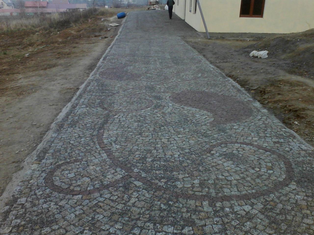 Wyjątkowy chodnik z kostki granitowej z dekoracyjnym wzorem ułożonym z ciemniejszego kamienia. Widok z perspektywy wzdłuż ścieżki, budynek w tle.