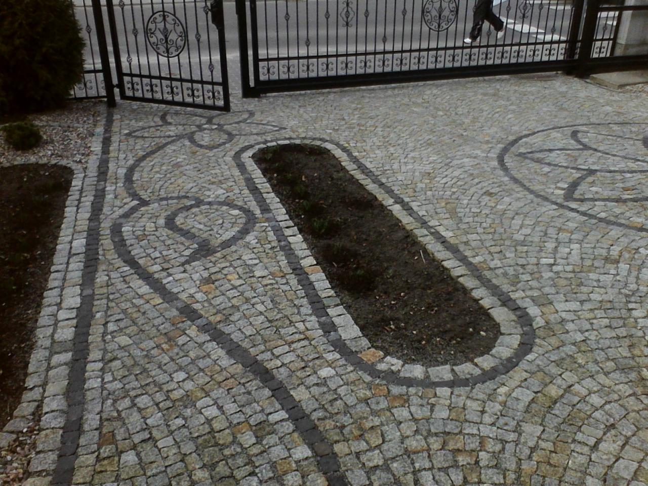Dekoracyjna nawierzchnia z kostki granitowej w odcieniach szarości i beżu, z wzorami kwiatowymi i podłużną rabatą, prowadząca do metalowej bramy.