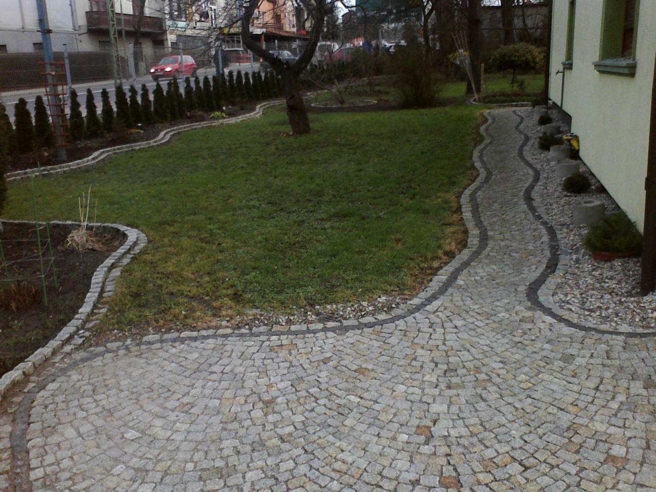Ścieżka z kostki granitowej w ogrodzie, z obrzeżami z ciemniejszej kostki. Widok na trawnik i roślinność, w tle ulica z czerwonym samochodem.