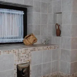 Jasne, kwadratowe płytki w łazience z oknem z firanką, niszami i dekoracjami. Mozaikowe wykończenia dodają rustykalnego charakteru wnętrzu.