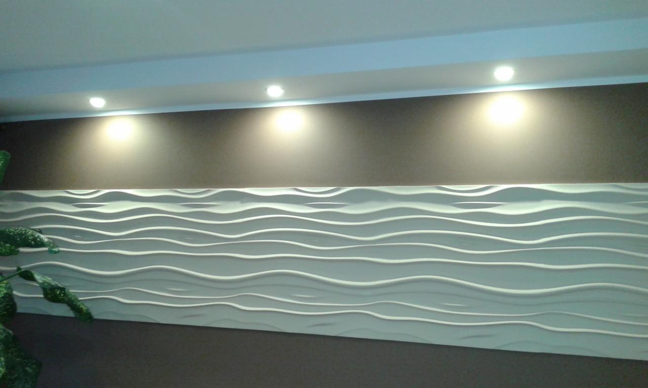 Panel ścienny 3D o falistym wzorze, oświetlony punktowo, w nowoczesnym wnętrzu. Fragment rośliny w lewym dolnym rogu kadru.  Dekoracyjne wykończenie ściany.