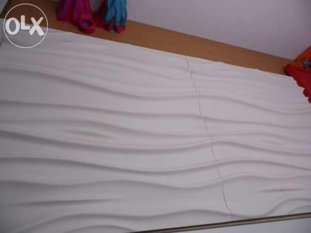 Białe panele ścienne 3D z delikatnym wzorem falistym, zamontowane w Pruszczu. U góry widoczny fragment drewnianej ramy i kolorowe elementy dekoracyjne.