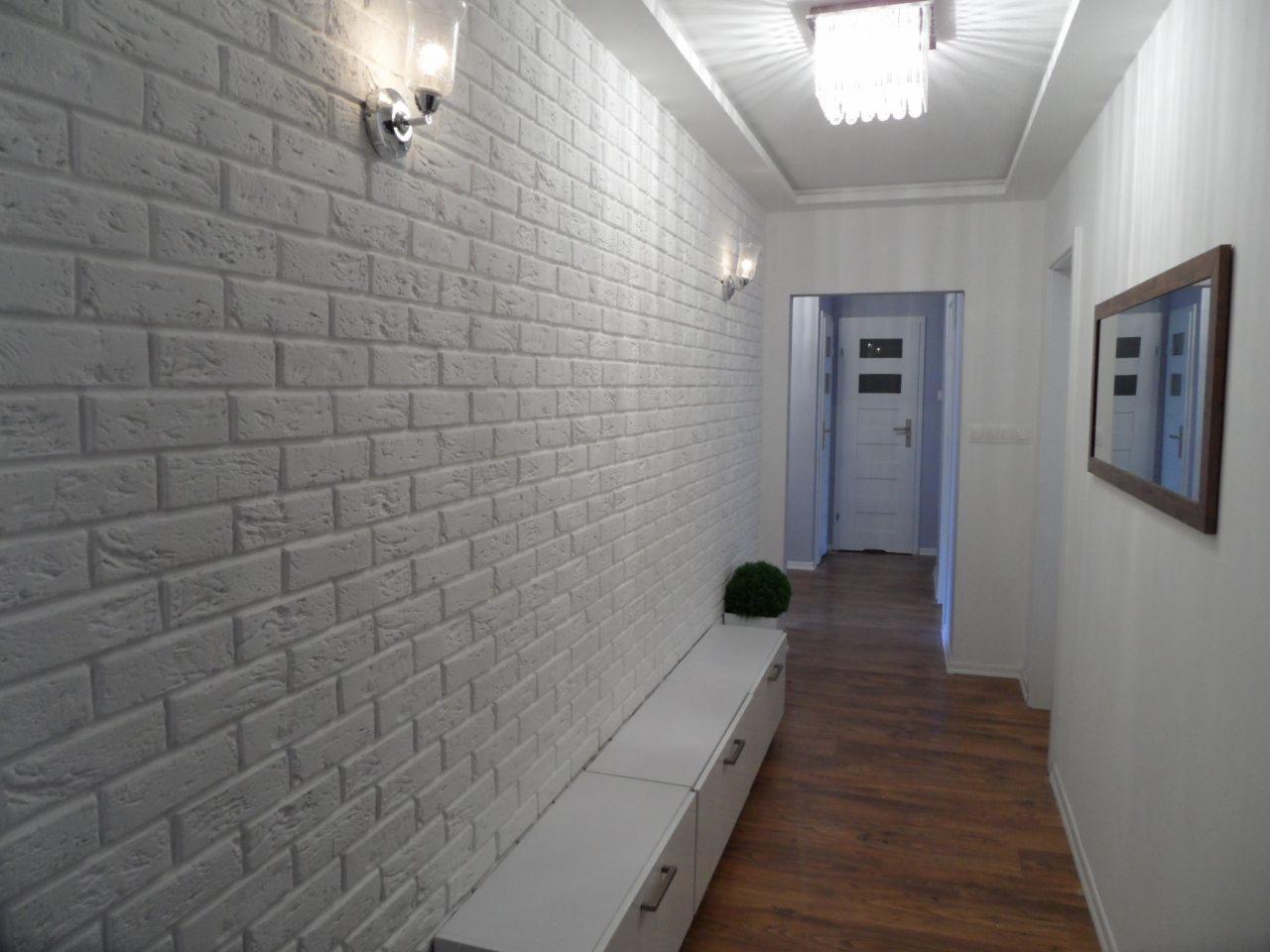 Wnętrze korytarza z białą cegłą na ścianie, oświetleniem punktowym i drewnianą podłogą. Lustro w brązowej ramie i minimalistyczna komoda dopełniają aranżację.