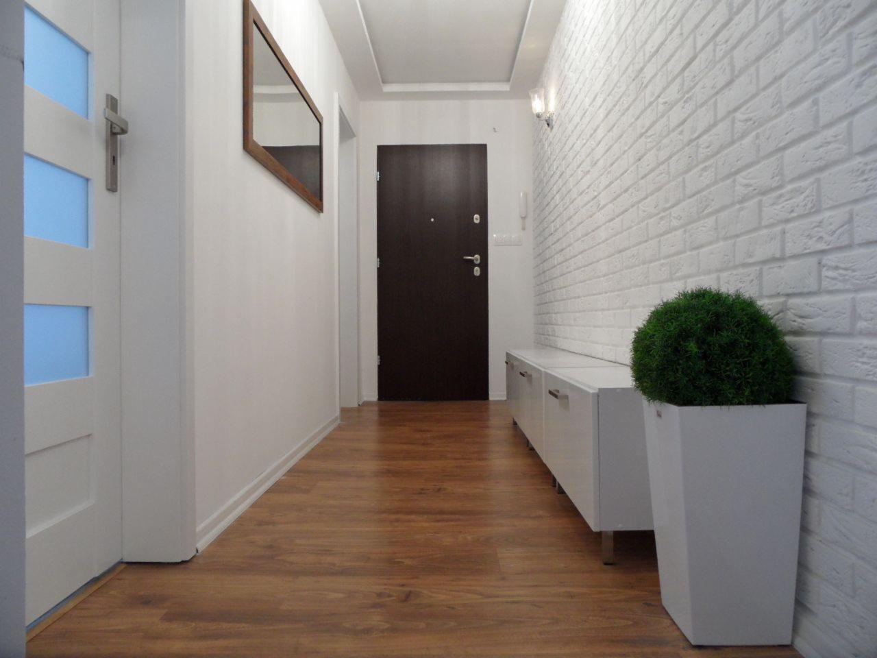 Nowoczesny przedpokój z białą cegłą na ścianie, drewnianą podłogą i minimalistycznym oświetleniem. Na ścianie lustro w drewnianej ramie, a obok drzwi roślina w donicy.