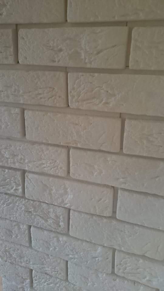 Ściana wyłożona dekoracyjną, białą cegłą z wyraźną fakturą, ułożoną w regularny wzór, z widocznymi spoinami. Ujęcie z bliska, lekko pod kątem.