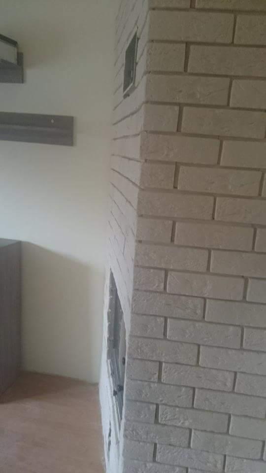 Szczegółowe ujęcie murowanej obudowy kominka z jasnej cegły, z widocznym otworem wentylacyjnym i fragmentem wnętrza pomieszczenia.