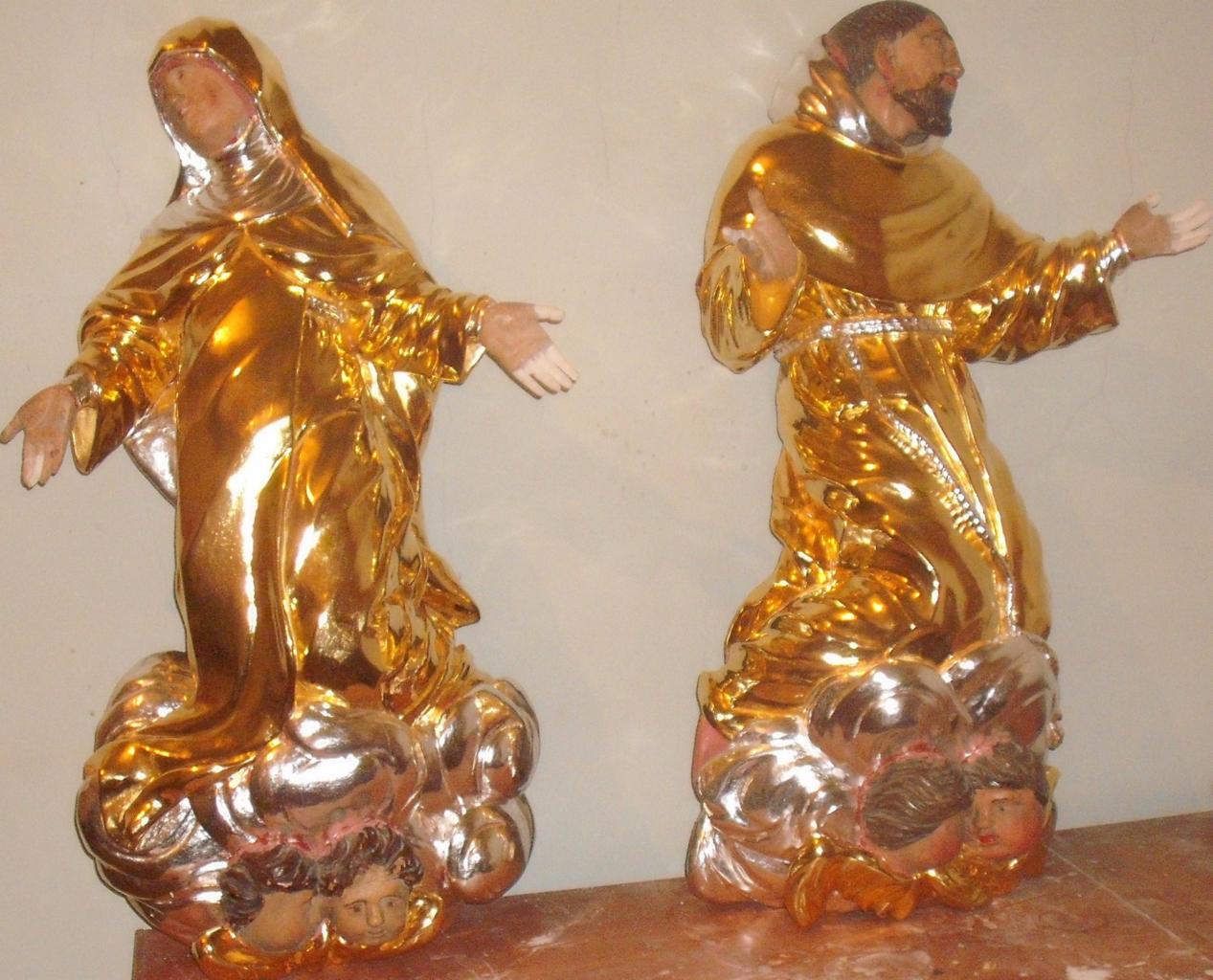 Dwie złote figury sakralne na marmurowym blacie. Figura kobiety z welonem i mężczyzny w habicie, obie na podstawie z aniołkami, oświetlone ciepłym światłem.