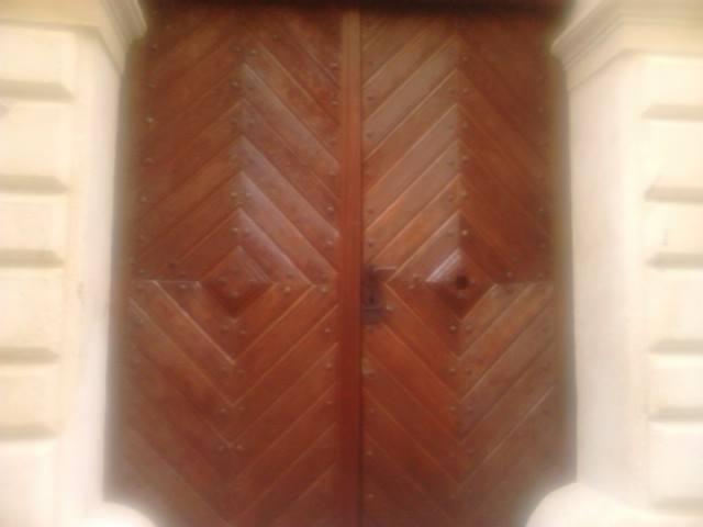 Drewniane, dwuskrzydłowe wrota z geometrycznym wzorem w jodełkę, osadzone w kamiennej fasadzie z dekoracyjnymi elementami architektonicznymi.