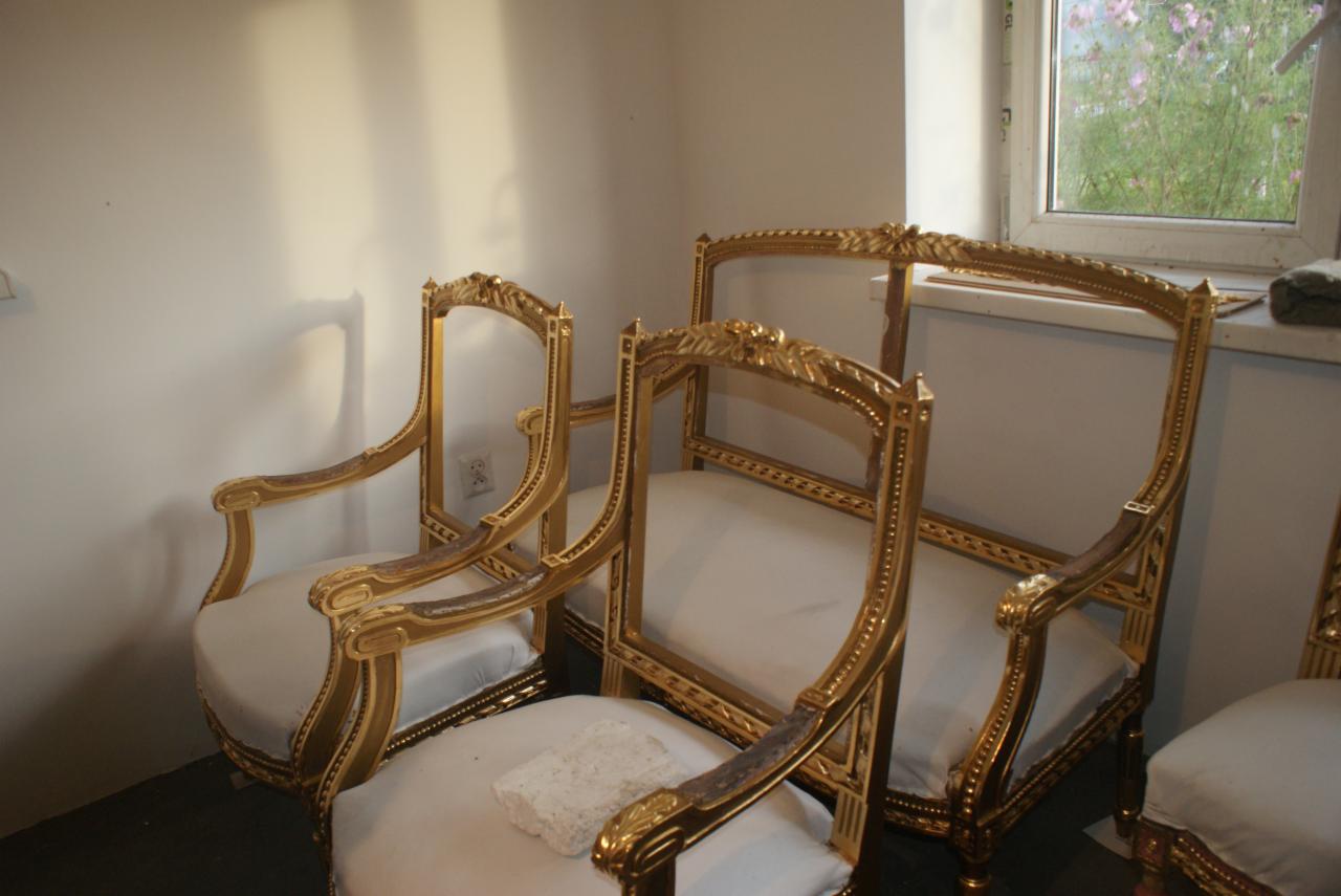 Zestaw odrestaurowanych, złoconych mebli w stylu Ludwika XVI: krzesła i kanapa z kremową tapicerką. Widoczne detale rzeźb i zdobień. W tle okno z widokiem na zieleń.