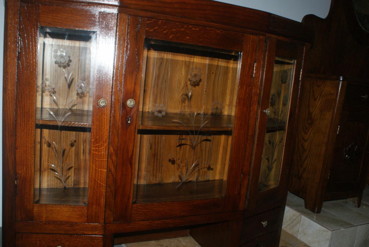 Ciemnobrązowy, drewniany kredens z przeszklonymi drzwiczkami zdobionymi kwiatowym motywem, widoczny z bliska, z elementami sąsiednich mebli w tle.