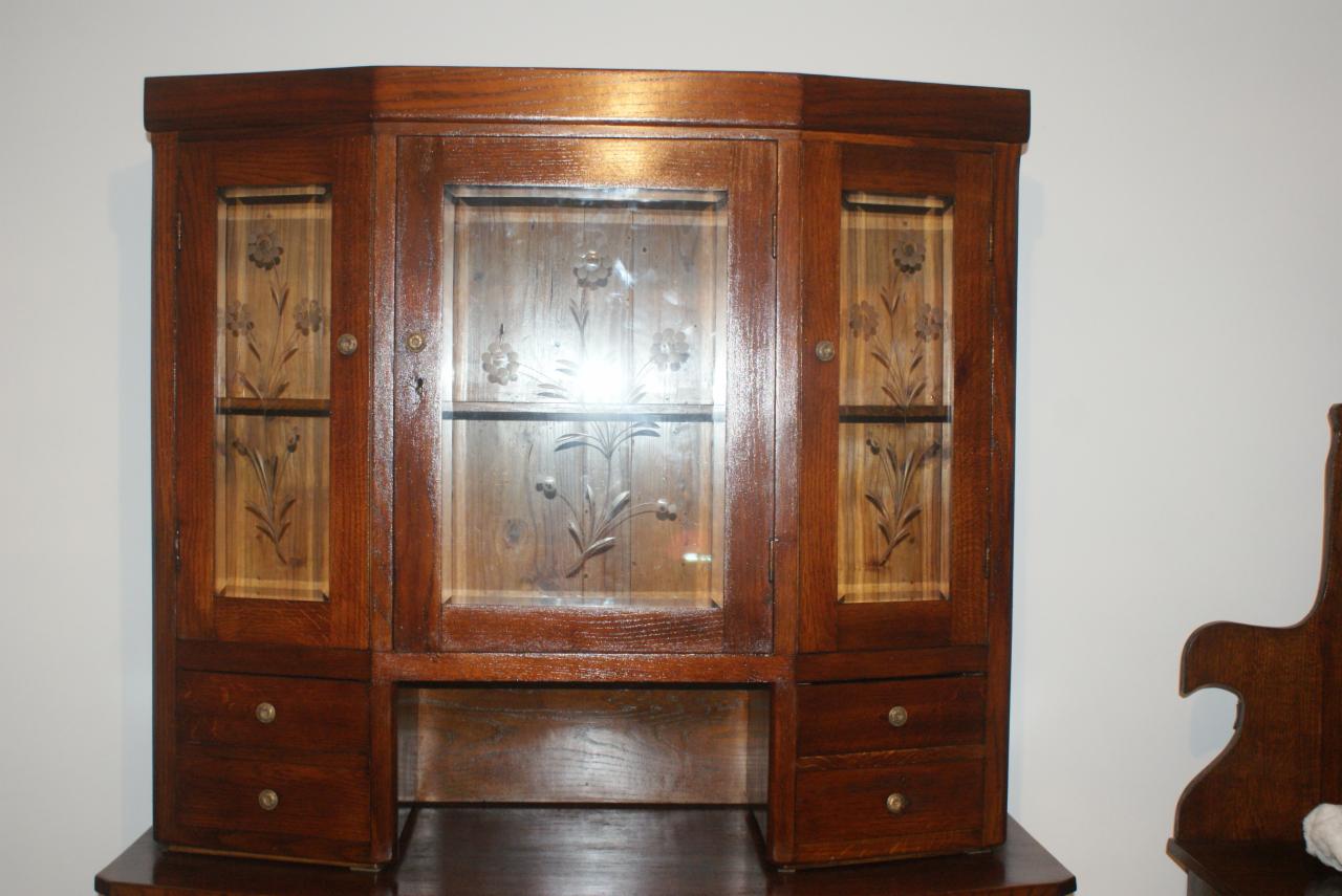 Kunsztowna, narożna szafka z drewna dębowego z przeszklonymi drzwiczkami, zdobiona motywem kwiatowym. Widoczne szuflady i delikatne detale wykończenia.