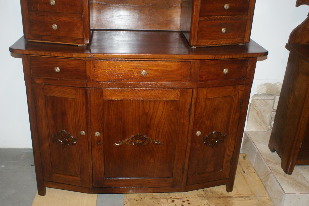 Antyczna, odnowiona komoda z ciemnego drewna z rzeźbionymi detalami, widoczna na tle jasnej ściany i fragmentu podłogi. Precyzyjna renowacja mebli.