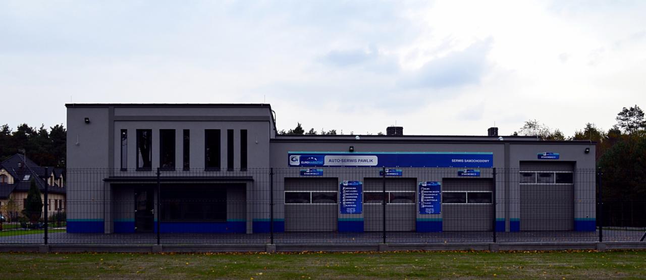 Szary budynek auto-serwisu Pawlik z niebieskimi akcentami, widoczny zza ogrodzenia. Na budynku szyld z nazwą i ofertą serwisu.