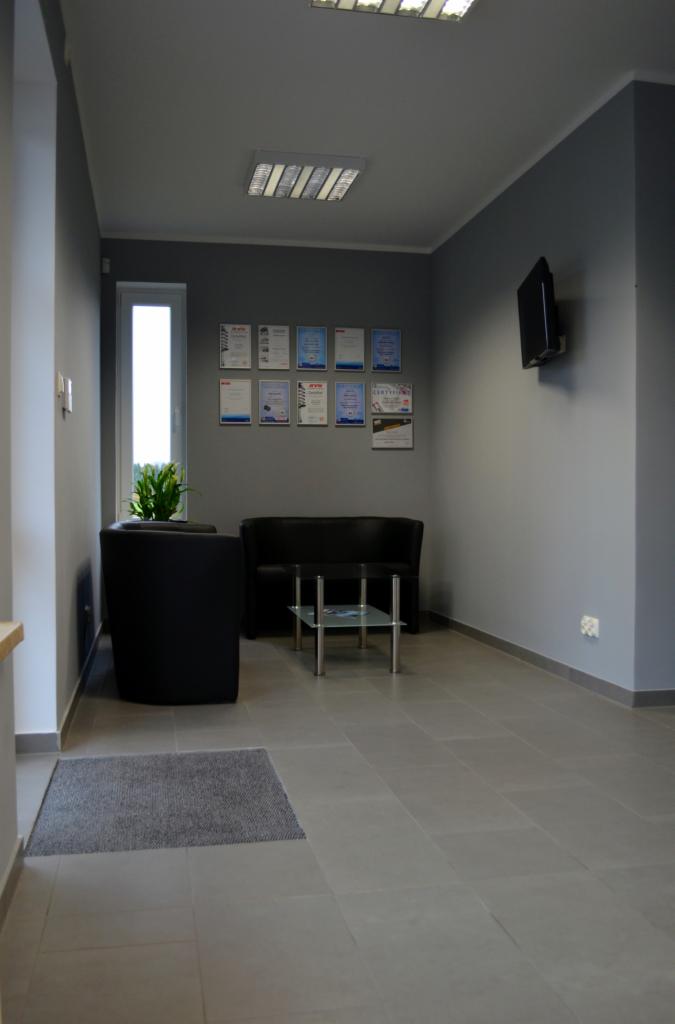 Poczekalnia warsztatu samochodowego z czarnymi sofami, szklanym stolikiem i ścianą z certyfikatami. Nowoczesne wnętrze w szarej tonacji.