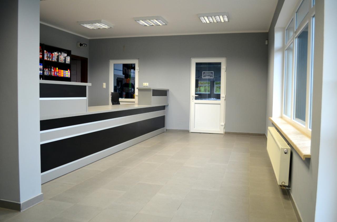 Wnętrze warsztatu samochodowego: recepcja z czarno-szarym blatem, okno z grzejnikiem, białe drzwi i regał z produktami. Stonowana kolorystyka, kafelki na podłodze.