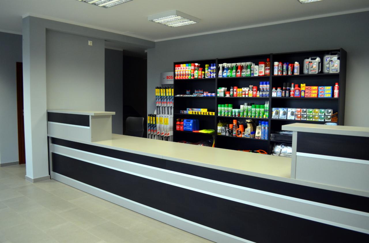 Wnętrze sklepu motoryzacyjnego: lada z czarno-szarymi pasami, półki z akcesoriami i płynami eksploatacyjnymi. Widoczny asortyment warsztatu.