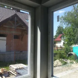 Wnętrze narożnego okna z widokiem na budynek w trakcie wznoszenia z cegły oraz dom z czerwonym dachem i zielenią.