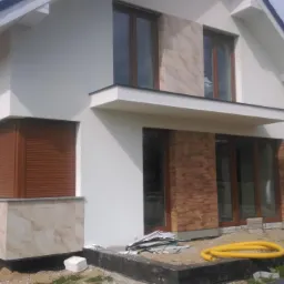 Elewacja domu jednorodzinnego z brązowymi roletami zewnętrznymi na parterze, wykończona białym tynkiem i panelami imitującymi cegłę, z widocznymi pozostałościami prac wykończeniowych na zewnątrz.