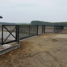 Nowoczesne ogrodzenie panelowe z bramą przesuwną i furtką w Garkach, z widoczną konstrukcją betonową u podstawy i polną drogą prowadzącą do posesji.