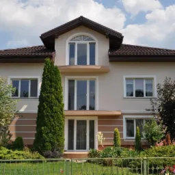 Dwupiętrowy dom z beżową elewacją, brązowym dachem i białymi ramami okiennymi, otoczony zielenią i niskim, zielonym płotem.