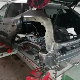 Szary samoch&oacute;d na podnośniku blacharskim z uszkodzonym tyłem, widoczne elementy konstrukcyjne nadwozia i narzędzia do naprawy.