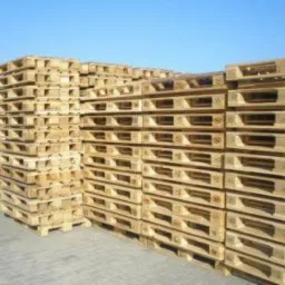 Stos palet drewnianych, ułożonych rzędami na zewnątrz, pod błękitnym niebem. Widoczne detale konstrukcji palet i faktura drewna. W tle zieleń drzew.