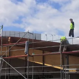Montaż blachy trapezowej na dachu budynku szkieletowego z użyciem rusztowania, dwóch pracowników w kamizelkach odblaskowych, widoczne panele izolacyjne.