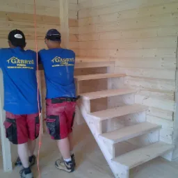 Wnętrze drewnianego domu w trakcie budowy. Widoczne drewniane schody, ściany z bali i dwóch pracowników firmy budowlanej w firmowych koszulkach.