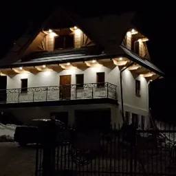 Dwukondygnacyjny dom z balkonem, oświetlony od dołu, sfotografowany nocą za ozdobnym metalowym ogrodzeniem. Widoczny fragment samochodu i zaspy śnieżne.