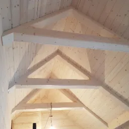 Surowa drewniana konstrukcja dachu z widocznymi belkami nośnymi i deskowaniem, jasne drewno, widok od wewnątrz.