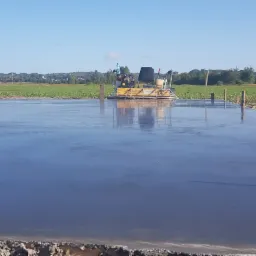 Świeżo wylana, gładka posadzka przemysłowa zacierana mechaniczną zacieraczką, widoczny błękitny samochód dostawczy w tle i drewniane kołki wyznaczające poziom.