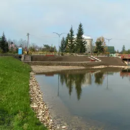 Widok na nowo wykopany staw z kamienistym brzegiem, trawą i schodami prowadzącymi do brzegu. W tle budynki, drzewa i czerwony samochód na małym moście.