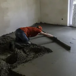 Pracownik w czerwonej koszulce wyrównuje szarą, mokrą wylewkę podłogową za pomocą długiej, drewnianej łaty; widoczne pryzmy nie rozprowadzonego materiału i płyty styropianowe o wzorze lastryko...