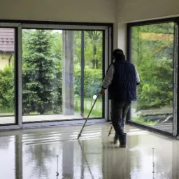 Pracownik rozprowadza mokrą wylewkę anhydrytową w przestronnym pomieszczeniu z dużymi oknami, widoczne bolce poziomujące w podłodze, za oknami zieleń i dom.