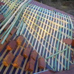 Układanie dachówki na dachu z drewnianą konstrukcją nośną. Widoczne elementy mocujące i przygotowane stosy dachówek.