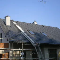 Dom w trakcie budowy z ciemną dachówką, oknami dachowymi i kominem. Aluminiowa drabina oparta o dach. Część budynku w surowym stanie.