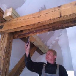 Bart- Bud - Pracownik maluje drewniane belki stropowe w pomieszczeniu z przygotowanymi ścianami, używając pędzla. Widoczne są charakterystyczne słoje drewna i detale konstrukcyjne.