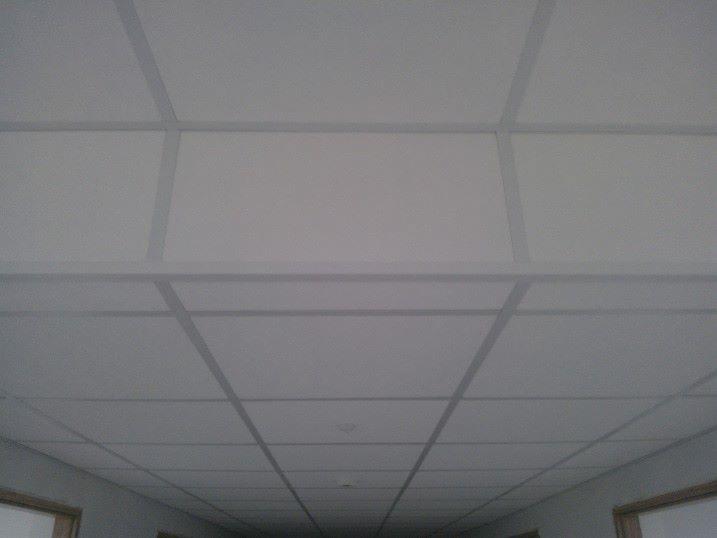 Kasetonowy sufit podwieszany w biurze, widok z dołu. Szare profile dzielą białe płyty. Perspektywa zbieżna w głąb korytarza.