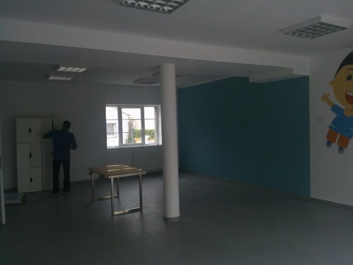 Świeżo pomalowane biuro we Wrocławiu z turkusową ścianą i postacią z bajki. Pracownik przy szafkach. Duże okno i kolumna na środku pomieszczenia.