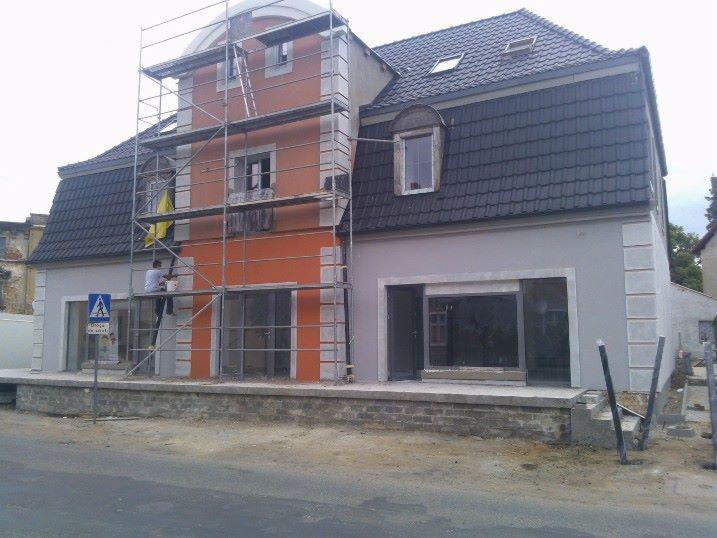 Prace elewacyjne: malowanie budynku z rusztowania. Część fasady pomalowana na pomarańczowo, druga w szarości. Widoczny znak drogowy i fragment ulicy.