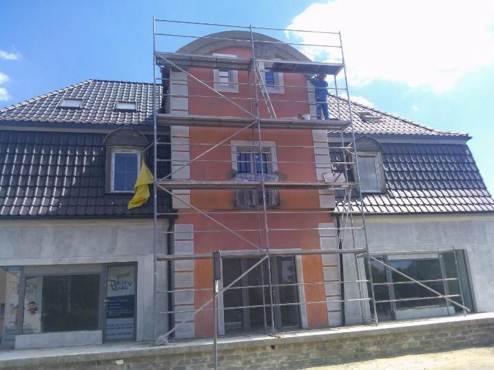 Pracownik w kombinezonie maluje elewację budynku z rusztowania. Widoczna odnowiona fasada z elementami dekoracyjnymi i fragment dachu z oknami.