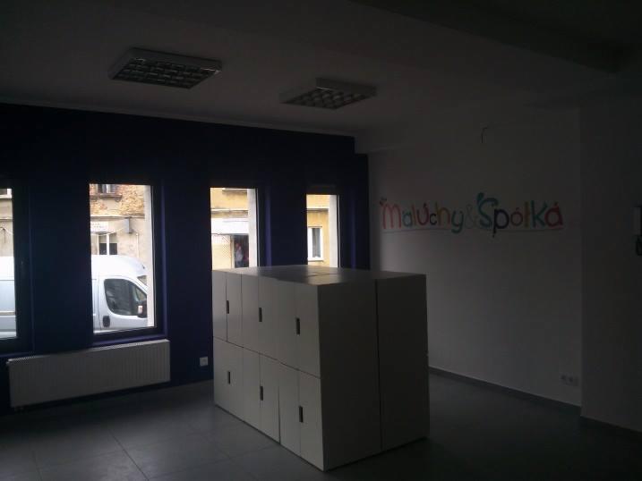 Wnętrze biura z szafkami na środku, okna z widokiem na ulicę, ściana z kolorowym napisem 'Maluchy & Spółka'.