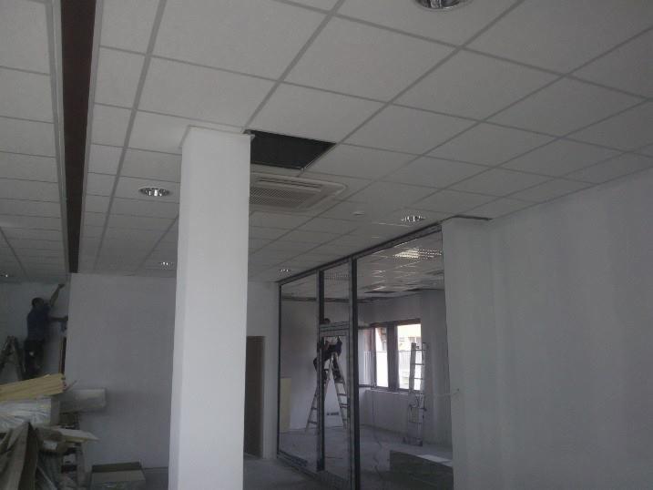 Malowanie ścian w biurze z kasetonowym sufitem podwieszanym. Widoczny malarz na drabinie i przeszklone ściany działowe. W tle okno.