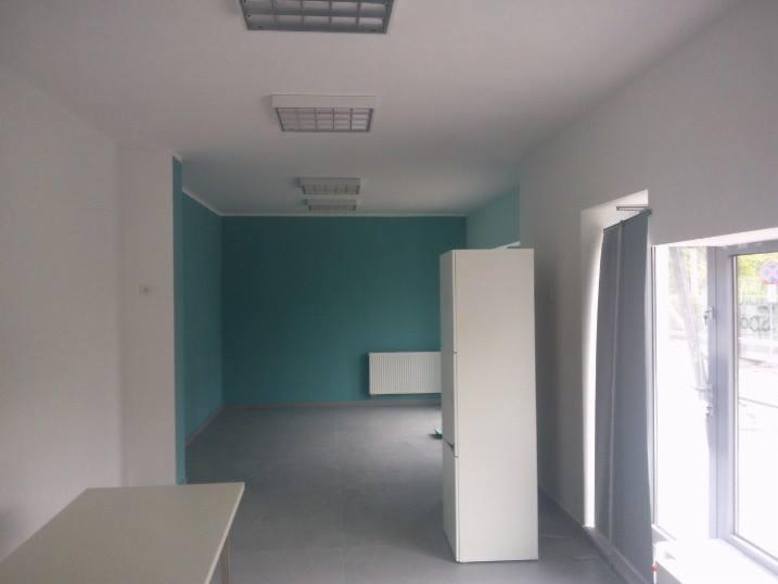 Pomalowane biuro z turkusową ścianą i białym grzejnikiem. Widoczne białe meble i okno z zasłoną. Oświetlenie sufitowe. Świeżo odmalowane ściany w biurze.