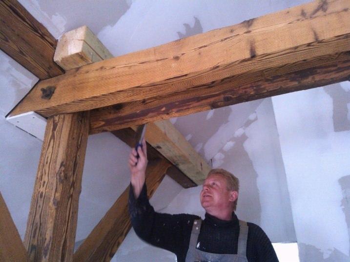 Pracownik maluje drewniane belki stropowe w pomieszczeniu z przygotowanymi ścianami, używając pędzla. Widoczne są charakterystyczne słoje drewna i detale konstrukcyjne.