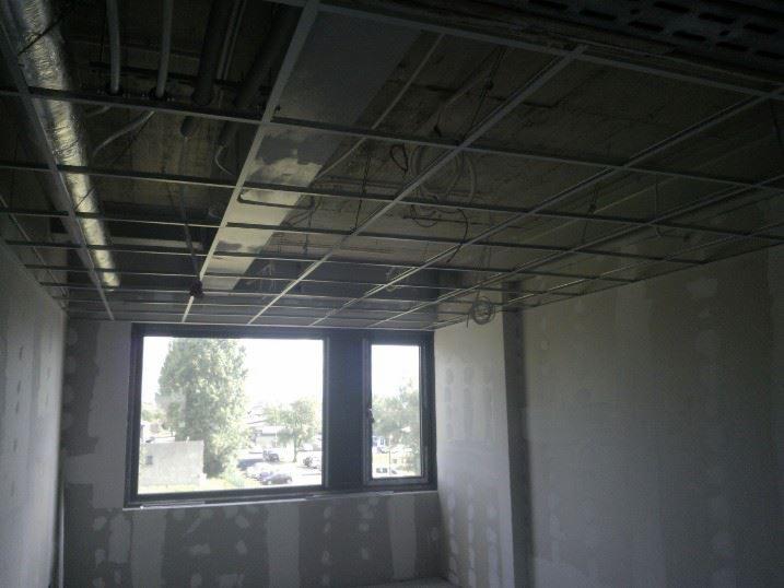 Wnętrze w trakcie prac wykończeniowych: sufit podwieszany na metalowej konstrukcji, ściany przygotowane do malowania. Widoczne okno z widokiem na zieleń.