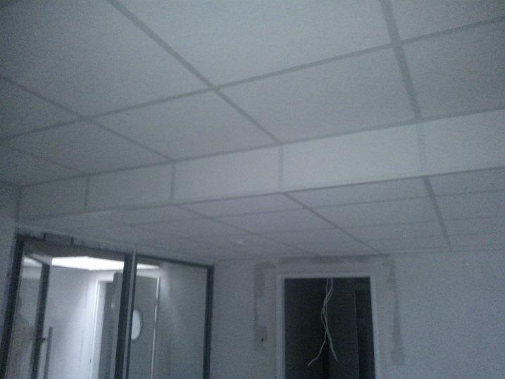 Kwadratowy sufit podwieszany w biurze, z widocznymi drzwiami i oknem, w trakcie prac wykończeniowych. Szare odcienie dominują w przestrzeni.