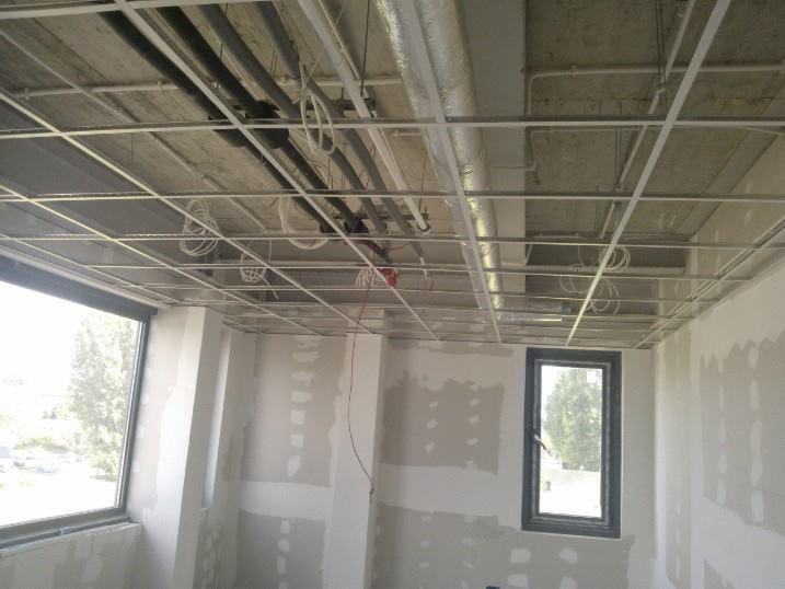 Wnętrze w trakcie prac wykończeniowych: widoczne profile sufitowe, instalacje pod stropem oraz ściany przygotowane do malowania z oknami.