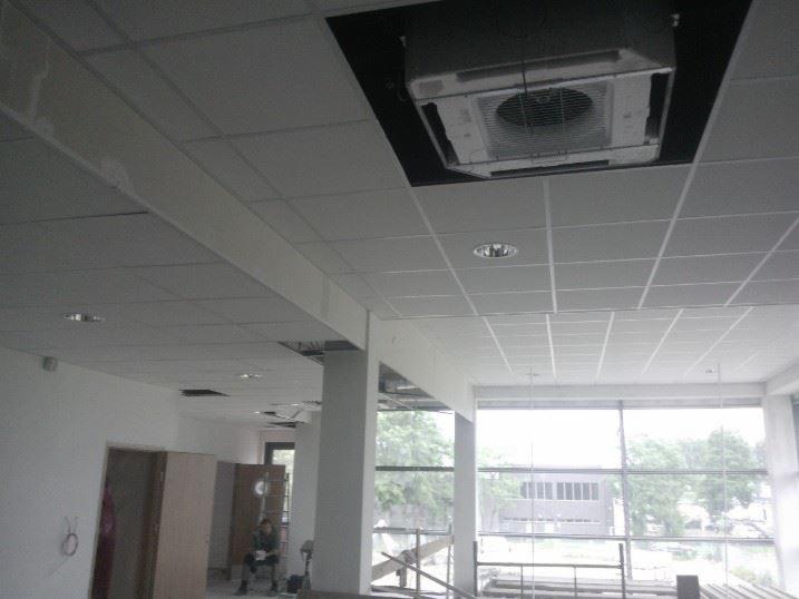 Sufit podwieszany kasetonowy z klimatyzacją w jasnym wnętrzu biurowym, widok na okna i filar, w tle sylwetka osoby. Prace wykończeniowe w trakcie.