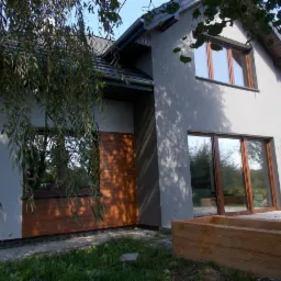 Szary dom jednorodzinny z drewnianą okładziną, widok z ogrodu, z elementami nowej elewacji i drewnianą donicą na pierwszym planie, częściowo zasłonięty przez zwisające gałęzie drzewa.